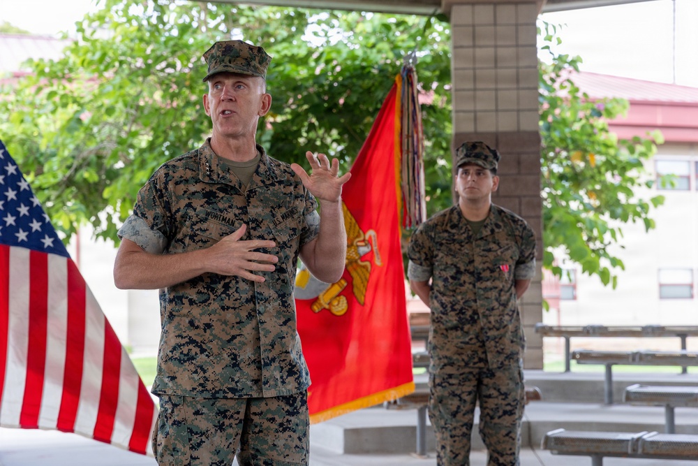 A U.S. Marine Corps officer, identified by nametape as 'WORTMAN,' is addressing an audience at what appears to be an outdoor military ceremony or event. He is gesturing with his hands while speaking, with a U.S. Marine Corps flag and an American flag visible in the background. A second Marine stands at ease behind him near the flags.