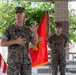 A U.S. Marine Corps officer, identified by nametape as 'WORTMAN,' is addressing an audience at what appears to be an outdoor military ceremony or event. He is gesturing with his hands while speaking, with a U.S. Marine Corps flag and an American flag visible in the background. A second Marine stands at ease behind him near the flags.