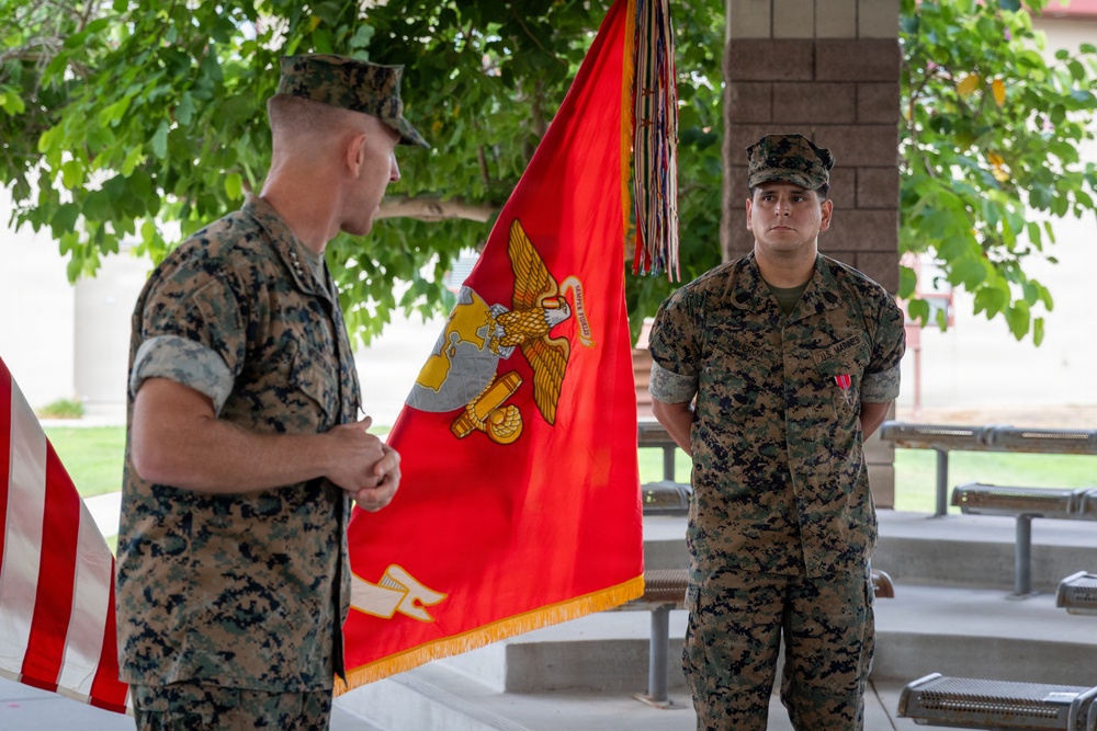Two U.S. Marines in woodland MARPAT camouflage uniforms are participating in a formal military ceremony outdoors. A senior Marine (facing away) appears to be addressing or presenting to a junior Marine who is standing at attention with a medal visible on his chest. A red Marine Corps unit guidon/colors flag with gold fringe is prominently displayed between them.
