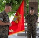 Two U.S. Marines in woodland MARPAT camouflage uniforms are participating in a formal military ceremony outdoors. A senior Marine (facing away) appears to be addressing or presenting to a junior Marine who is standing at attention with a medal visible on his chest. A red Marine Corps unit guidon/colors flag with gold fringe is prominently displayed between them.