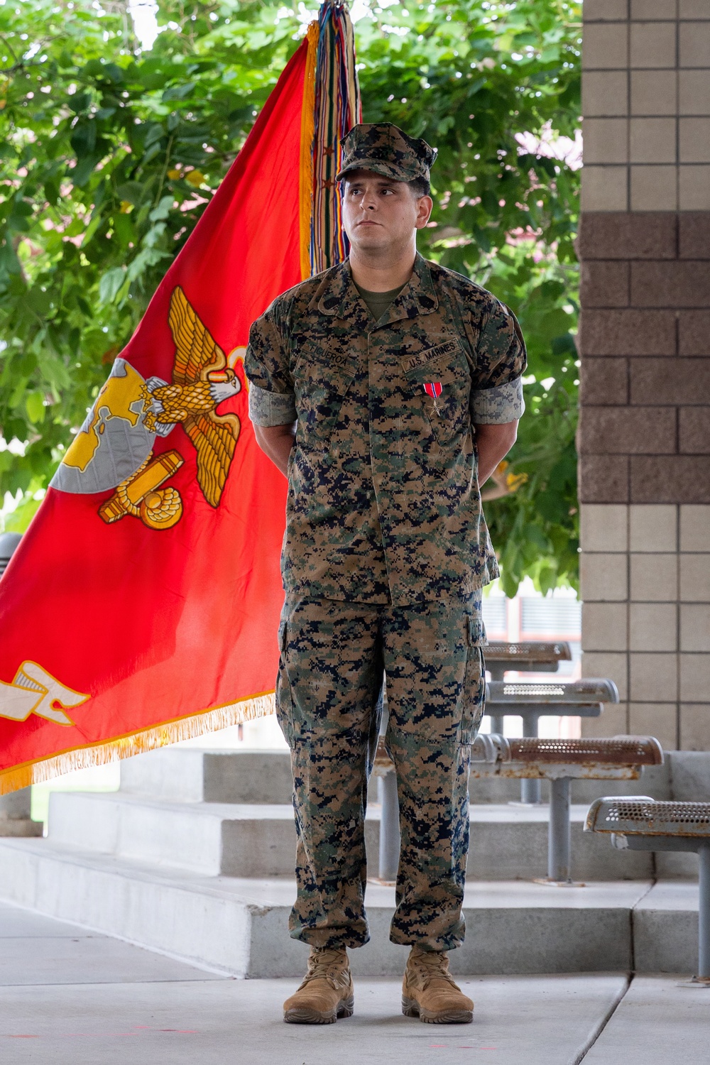 A U.S. Marine stands at attention in MARPAT woodland camouflage uniform in front of a red Marine Corps organizational flag (guidon). The Marine is wearing a single medal on his chest and has his hands clasped behind his back, suggesting a formal military ceremony or award presentation. The setting appears to be an outdoor plaza or courtyard with steps, benches, and trees visible in the background.