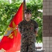 A U.S. Marine stands at attention in MARPAT woodland camouflage uniform in front of a red Marine Corps organizational flag (guidon). The Marine is wearing a single medal on his chest and has his hands clasped behind his back, suggesting a formal military ceremony or award presentation. The setting appears to be an outdoor plaza or courtyard with steps, benches, and trees visible in the background.