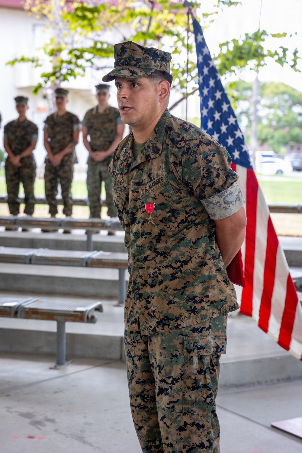 A U.S. Marine in MARPAT digital camouflage uniform stands at the forefront of what appears to be a military award ceremony. He is wearing a medal on his chest, consistent with a Bronze Star Medal with 'V' device, and has a 'U.S. MARINES' tape visible on his uniform. An American flag is positioned behind him, and several other Marines stand at ease in the background.
