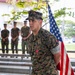 A U.S. Marine in MARPAT digital camouflage uniform stands at the forefront of what appears to be a military award ceremony. He is wearing a medal on his chest, consistent with a Bronze Star Medal with 'V' device, and has a 'U.S. MARINES' tape visible on his uniform. An American flag is positioned behind him, and several other Marines stand at ease in the background.