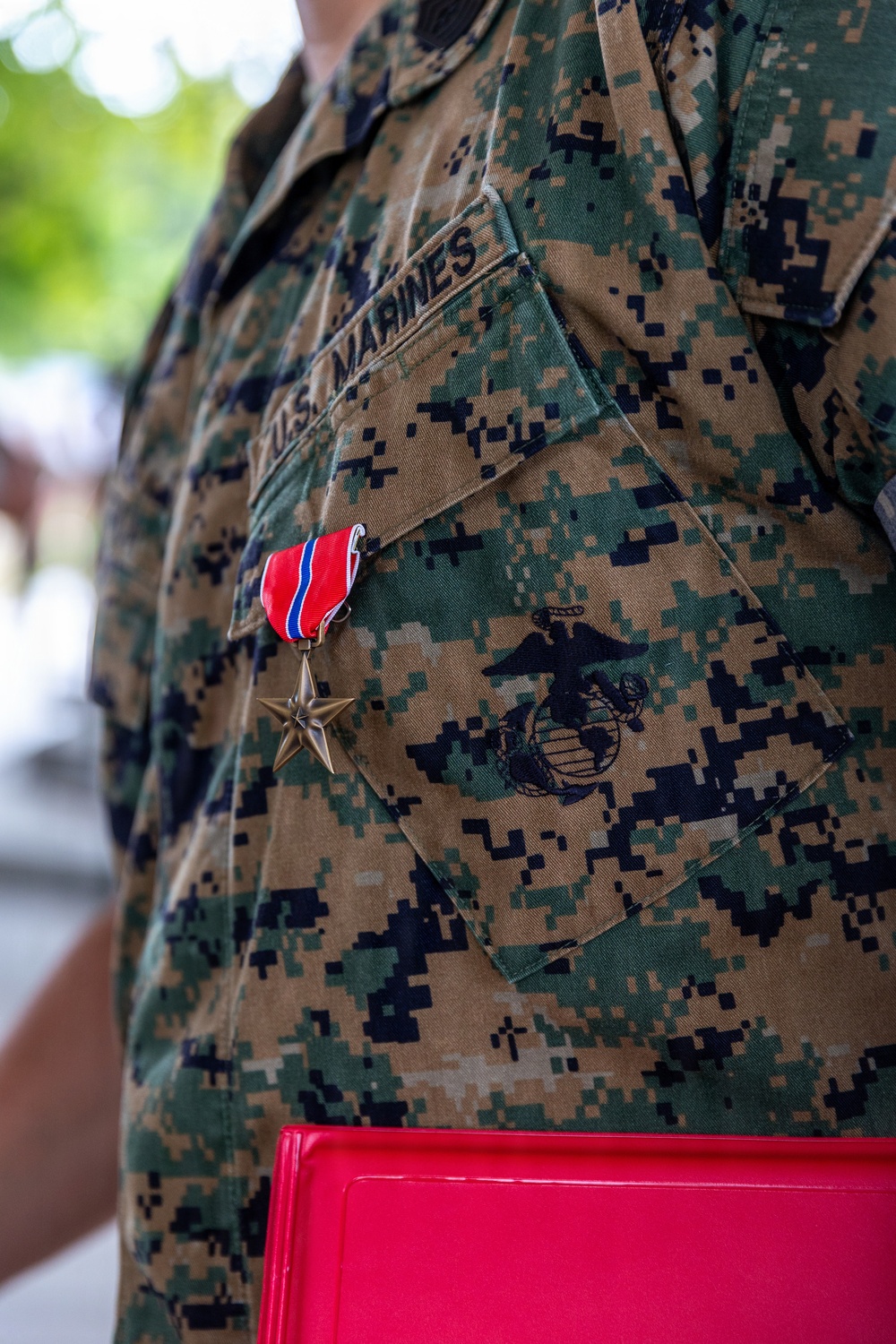 A close-up photograph of a U.S. Marines service member wearing the MARPAT (Marine Pattern) digital camouflage uniform (MCCUU). A Bronze Star Medal with ribbon is pinned to the left chest pocket, and the Eagle, Globe, and Anchor (EGA) emblem is embroidered on the pocket flap. The individual is holding a red folder or binder at their side.
