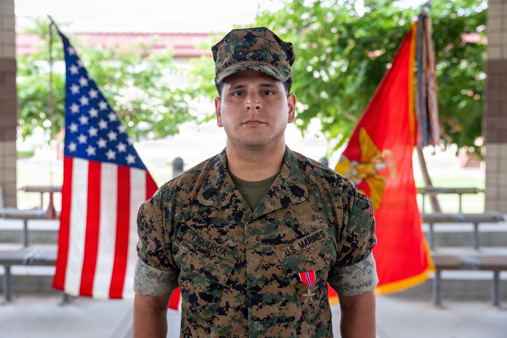 A U.S. Marine identified by name tape as 'FIGUEROA' stands at attention in Marine Pattern (MARPAT) woodland camouflage uniform. He is wearing a Bronze Star Medal with Valor device on his chest, indicating a formal award ceremony. An American flag and a U.S. Marine Corps flag are visible in the background.