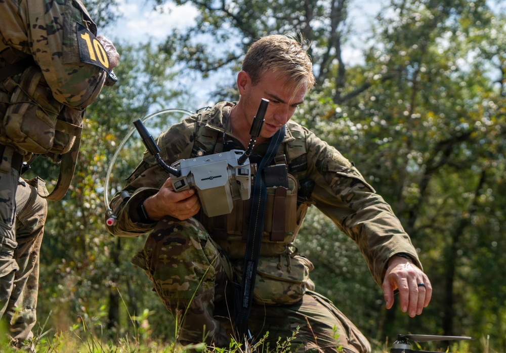 A soldier in multicam camouflage uniform is operating what appears to be a DJI drone controller in an outdoor wooded setting. He is crouching low to the ground, focused on the controller, while a second soldier wearing a number '10' identifier patch is partially visible to the left. The scene appears to be a military training exercise involving unmanned aerial vehicle (UAV) operations.