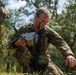A soldier in multicam camouflage uniform is operating what appears to be a DJI drone controller in an outdoor wooded setting. He is crouching low to the ground, focused on the controller, while a second soldier wearing a number '10' identifier patch is partially visible to the left. The scene appears to be a military training exercise involving unmanned aerial vehicle (UAV) operations.