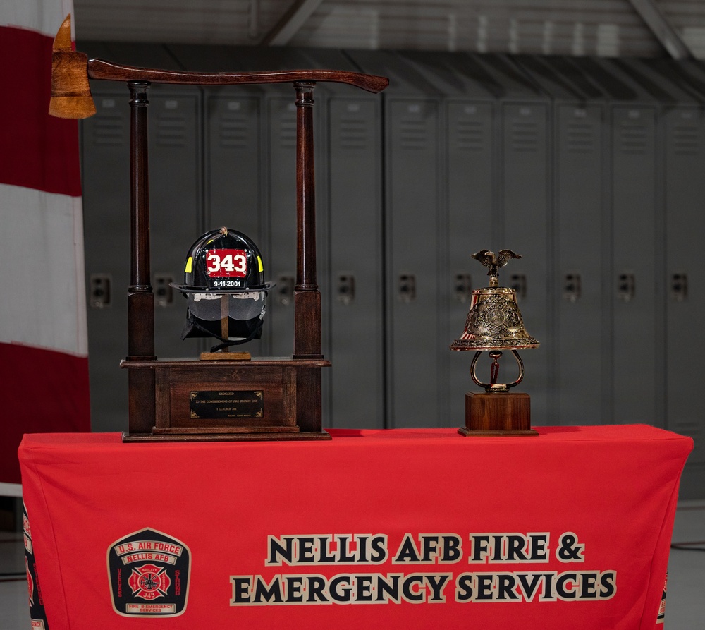 A memorial display at Nellis AFB Fire &amp; Emergency Services features a firefighter's helmet marked '343' and '9-11-2001' mounted on a wooden stand with a fire axe, commemorating the 343 FDNY firefighters lost on September 11, 2001. A decorative brass bell on a wooden base sits alongside the helmet display. Both items rest on a red tablecloth bearing the Nellis AFB Fire &amp; Emergency Services insignia and text.