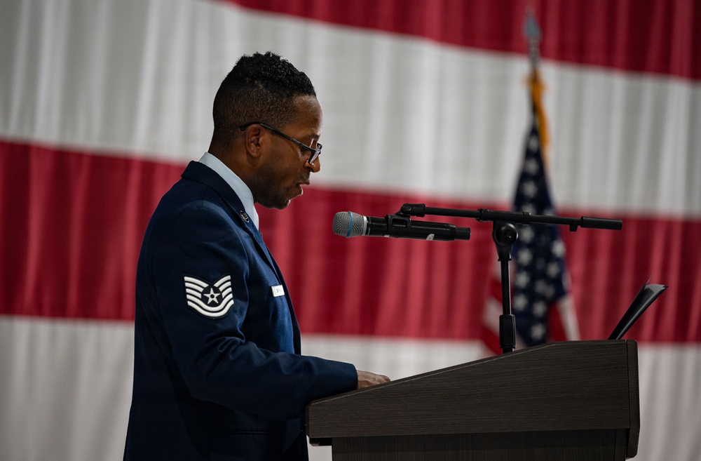 A U.S. Air Force Technical Sergeant stands at a podium and speaks into a microphone at what appears to be an official military ceremony. He is wearing the Air Force Service Dress uniform with visible rank insignia on his sleeve. A large American flag and a U.S. flag on a stand are visible in the background.