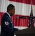 A U.S. Air Force Technical Sergeant stands at a podium and speaks into a microphone at what appears to be an official military ceremony. He is wearing the Air Force Service Dress uniform with visible rank insignia on his sleeve. A large American flag and a U.S. flag on a stand are visible in the background.