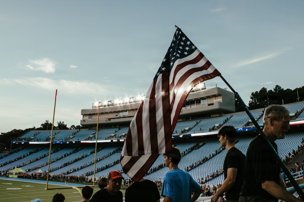 An American flag is being carried or held at field level inside Kenan Stadium, a college football venue. The scene appears to be during a pre-game or ceremonial event, with stadium lights illuminating the press box area against a late afternoon sky. The stands are partially filled with spectators visible in the background.