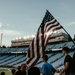 An American flag is being carried or held at field level inside Kenan Stadium, a college football venue. The scene appears to be during a pre-game or ceremonial event, with stadium lights illuminating the press box area against a late afternoon sky. The stands are partially filled with spectators visible in the background.