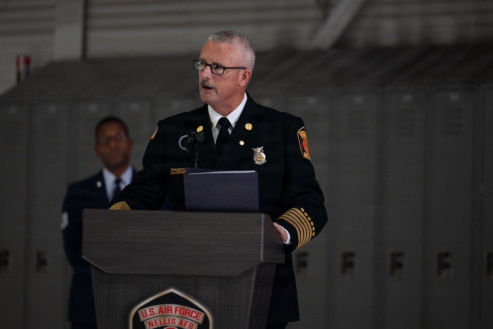 A senior fire department official in formal dress uniform is speaking at a podium bearing the U.S. Air Force Nellis AFB insignia. The event appears to be a formal ceremony held inside what looks like an aircraft hangar, with metal lockers visible in the background. A uniformed Air Force member is visible standing in the background.