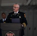 A senior fire department official in formal dress uniform is speaking at a podium bearing the U.S. Air Force Nellis AFB insignia. The event appears to be a formal ceremony held inside what looks like an aircraft hangar, with metal lockers visible in the background. A uniformed Air Force member is visible standing in the background.
