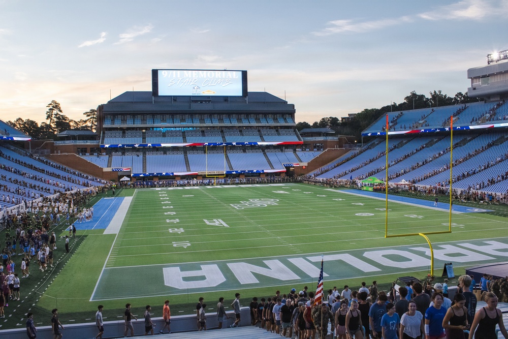 The image shows Kenan Memorial Stadium at the University of North Carolina at Chapel Hill during a 9/11 Memorial Stair Climb event. The field, marked with 'CAROLINA' in the end zone, is surrounded by crowds of participants on the track and sidelines. The event is taking place at dusk, with the scoreboard prominently displaying '9/11 Memorial Stair Climb'.