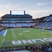 The image shows Kenan Memorial Stadium at the University of North Carolina at Chapel Hill during a 9/11 Memorial Stair Climb event. The field, marked with 'CAROLINA' in the end zone, is surrounded by crowds of participants on the track and sidelines. The event is taking place at dusk, with the scoreboard prominently displaying '9/11 Memorial Stair Climb'.