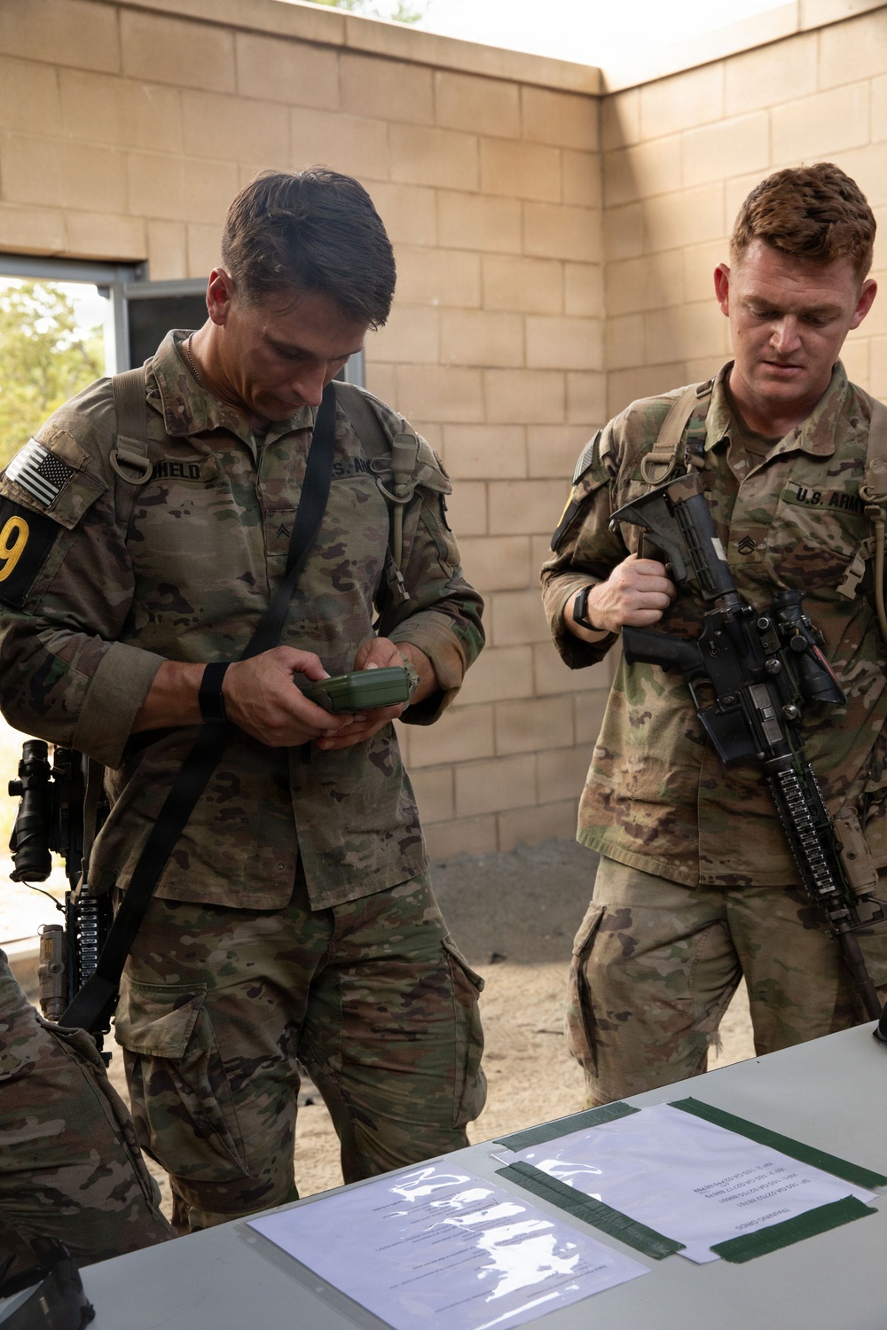 Two U.S. Army soldiers in MultiCam/OCP camouflage uniforms are standing at a table outdoors near a cinder block structure. The soldier on the left is examining a small green object in his hands, while the soldier on the right is holding an M4-style carbine rifle. Documents and papers are visible on the table in front of them, suggesting a briefing or administrative checkpoint.