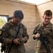 Two U.S. Army soldiers in MultiCam/OCP camouflage uniforms are standing at a table outdoors near a cinder block structure. The soldier on the left is examining a small green object in his hands, while the soldier on the right is holding an M4-style carbine rifle. Documents and papers are visible on the table in front of them, suggesting a briefing or administrative checkpoint.