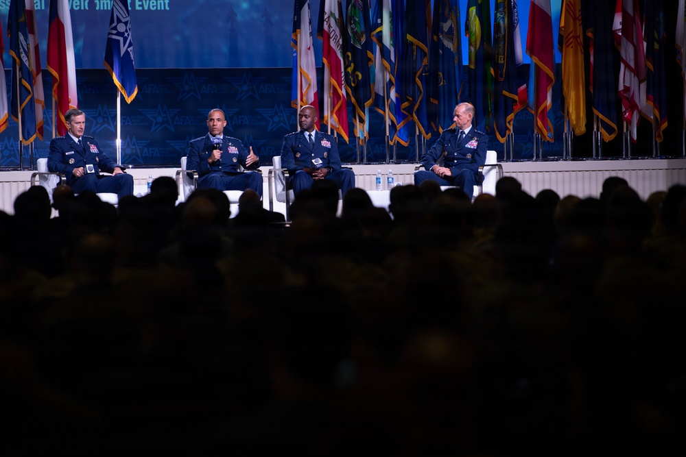Four military officers in dress blue uniforms are seated on a stage panel at what appears to be an Air Force Association (AFA) event, as indicated by the AFA logos visible on the backdrop. One officer is speaking into a microphone while the others listen. A large audience is visible in silhouette in the foreground.