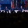 Four military officers in dress blue uniforms are seated on a stage panel at what appears to be an Air Force Association (AFA) event, as indicated by the AFA logos visible on the backdrop. One officer is speaking into a microphone while the others listen. A large audience is visible in silhouette in the foreground.