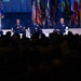 Four military officers in dress blue uniforms are seated on a stage panel at what appears to be an Air Force Association (AFA) event, as indicated by the AFA logos visible on the backdrop. One officer is speaking into a microphone while the others listen. A large audience is visible in silhouette in the foreground.