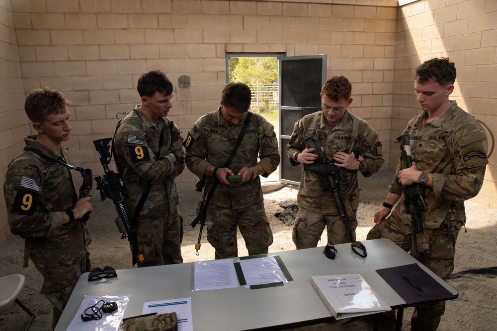 Five U.S. Army soldiers in MultiCam/OCP camouflage uniforms are gathered around a table inside a cinder block building, appearing to review documents and equipment. Several soldiers are holding M4/M16-style rifles, and at least two are wearing the number '9' on their uniforms, suggesting a competition or evaluation event. A document on the table is partially visible and appears to reference a competition.