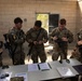 Five U.S. Army soldiers in MultiCam/OCP camouflage uniforms are gathered around a table inside a cinder block building, appearing to review documents and equipment. Several soldiers are holding M4/M16-style rifles, and at least two are wearing the number '9' on their uniforms, suggesting a competition or evaluation event. A document on the table is partially visible and appears to reference a competition.