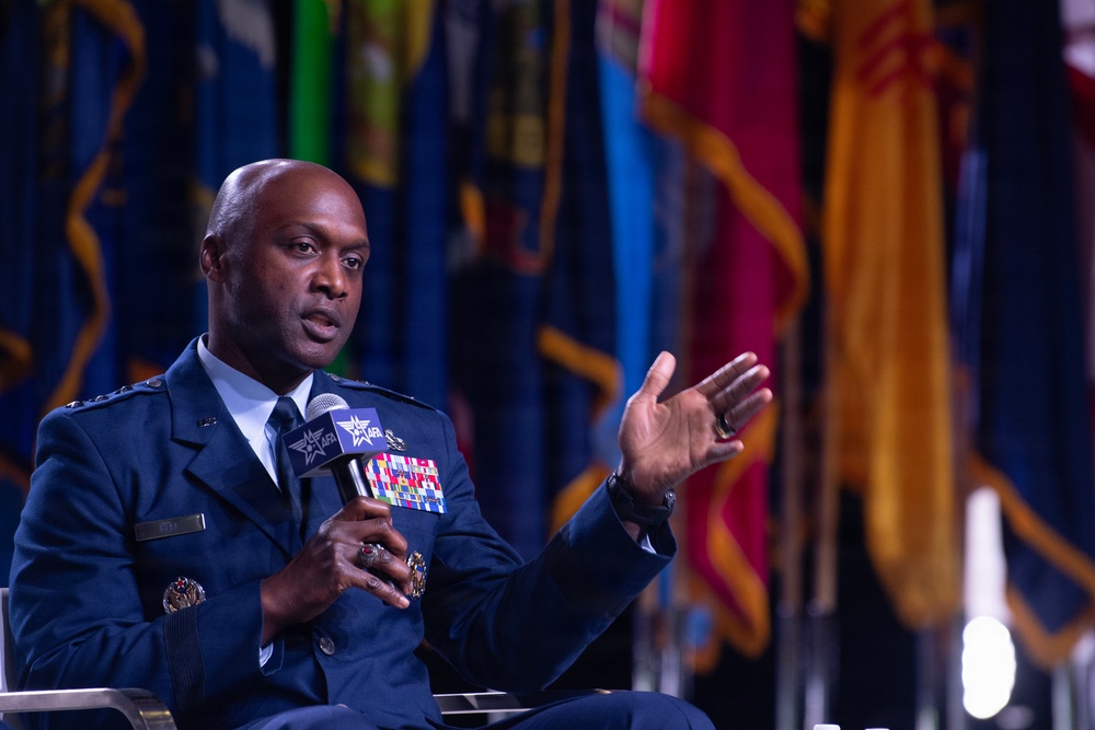 A high-ranking military officer in a dark blue dress uniform is seated and speaking into a microphone branded with the AFA (Air Force Association) logo. He is gesturing with his right hand, suggesting active engagement in a panel discussion or interview. Multiple colorful flags are visible in the blurred background, indicating a formal military or governmental event.