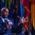 A high-ranking military officer in a dark blue dress uniform is seated and speaking into a microphone branded with the AFA (Air Force Association) logo. He is gesturing with his right hand, suggesting active engagement in a panel discussion or interview. Multiple colorful flags are visible in the blurred background, indicating a formal military or governmental event.