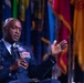 A high-ranking military officer in a dark blue dress uniform is seated and speaking into a microphone branded with the AFA (Air Force Association) logo. He is gesturing with his right hand, suggesting active engagement in a panel discussion or interview. Multiple colorful flags are visible in the blurred background, indicating a formal military or governmental event.