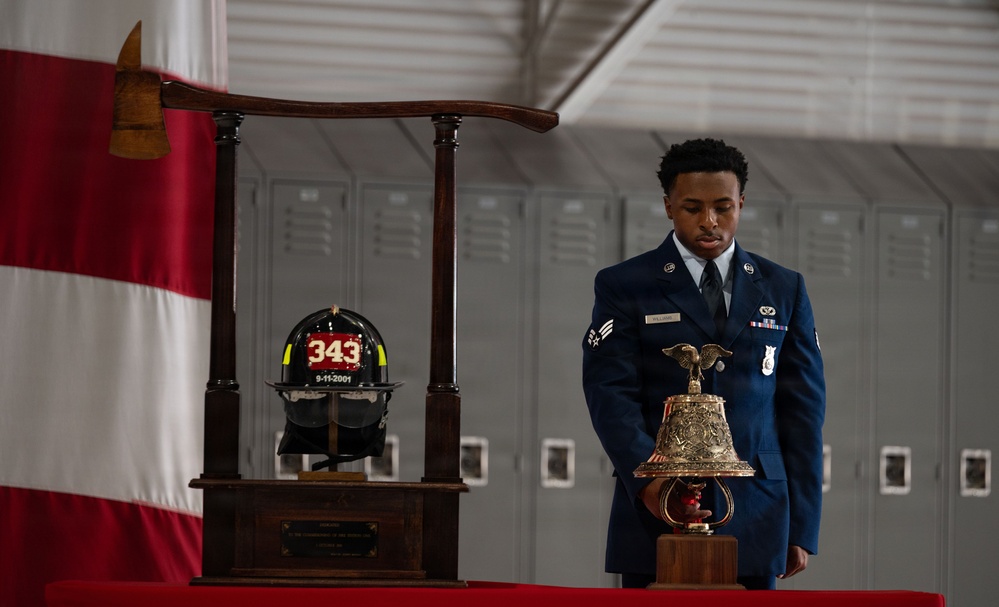A U.S. Air Force service member in dress blues stands solemnly at a 9/11 memorial display, holding a ceremonial brass bell. The display includes a firefighter helmet marked '343' and '9-11-2001', commemorating the 343 FDNY firefighters who died on September 11, 2001. The setting appears to be inside a military hangar or facility with lockers visible in the background.
