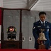 A U.S. Air Force service member in dress blues stands solemnly at a 9/11 memorial display, holding a ceremonial brass bell. The display includes a firefighter helmet marked '343' and '9-11-2001', commemorating the 343 FDNY firefighters who died on September 11, 2001. The setting appears to be inside a military hangar or facility with lockers visible in the background.