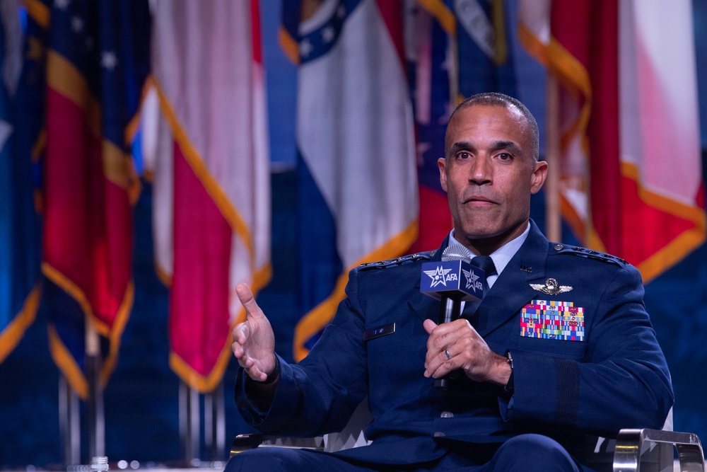 A senior military officer in a dark blue dress uniform is seated and speaking into an AFA (Air Force Association) branded microphone at what appears to be a formal conference or symposium. He is gesturing with his left hand while holding the microphone, suggesting active engagement with an audience. Multiple military and state flags are visible in the blurred background.