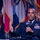 A senior military officer in a dark blue dress uniform is seated and speaking into an AFA (Air Force Association) branded microphone at what appears to be a formal conference or symposium. He is gesturing with his left hand while holding the microphone, suggesting active engagement with an audience. Multiple military and state flags are visible in the blurred background.