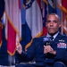 A senior military officer in a dark blue dress uniform is seated and speaking into an AFA (Air Force Association) branded microphone at what appears to be a formal conference or symposium. He is gesturing with his left hand while holding the microphone, suggesting active engagement with an audience. Multiple military and state flags are visible in the blurred background.