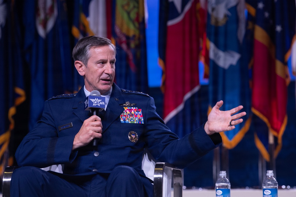 A senior U.S. Air Force officer in dress blue uniform is seated and speaking into an AFA (Air Force Association) branded microphone at what appears to be a formal military conference or symposium. The officer displays multiple rows of service ribbons, pilot wings, and four-star rank insignia on their uniform. Multiple U.S. state flags are visible in the background, suggesting a large official venue.