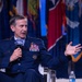 A senior U.S. Air Force officer in dress blue uniform is seated and speaking into an AFA (Air Force Association) branded microphone at what appears to be a formal military conference or symposium. The officer displays multiple rows of service ribbons, pilot wings, and four-star rank insignia on their uniform. Multiple U.S. state flags are visible in the background, suggesting a large official venue.