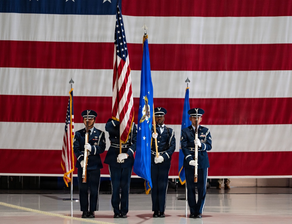 A U.S. Air Force color guard stands at attention during a formal military ceremony inside what appears to be a large hangar. Four service members in dress blue uniforms carry flags including the U.S. national flag and the U.S. Air Force flag. A large American flag serves as a dramatic backdrop behind the formation.