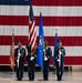A U.S. Air Force color guard stands at attention during a formal military ceremony inside what appears to be a large hangar. Four service members in dress blue uniforms carry flags including the U.S. national flag and the U.S. Air Force flag. A large American flag serves as a dramatic backdrop behind the formation.