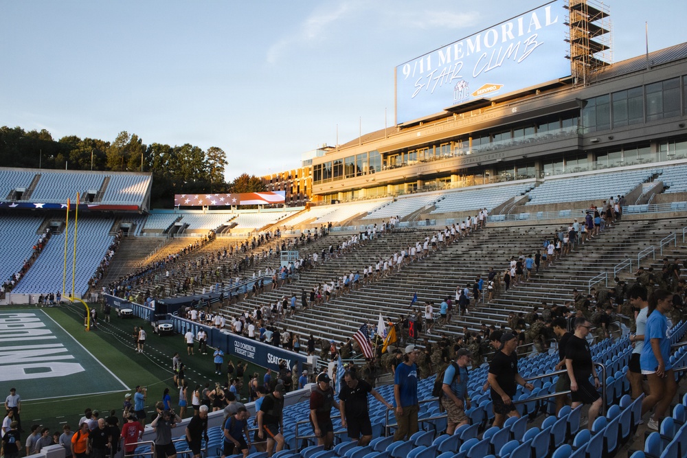 A large group of participants are climbing stadium stairs at what appears to be a college football stadium during a 9/11 Memorial Stair Climb event. The scoreboard prominently displays '9/11 MEMORIAL STAIR CLIMB' and participants, many in military attire and civilian clothing, are spread across multiple sections of the stadium bleachers. The event appears to be taking place during golden hour, with an American flag visible on the field level.