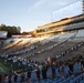 A large group of participants are climbing stadium stairs at what appears to be a college football stadium during a 9/11 Memorial Stair Climb event. The scoreboard prominently displays '9/11 MEMORIAL STAIR CLIMB' and participants, many in military attire and civilian clothing, are spread across multiple sections of the stadium bleachers. The event appears to be taking place during golden hour, with an American flag visible on the field level.