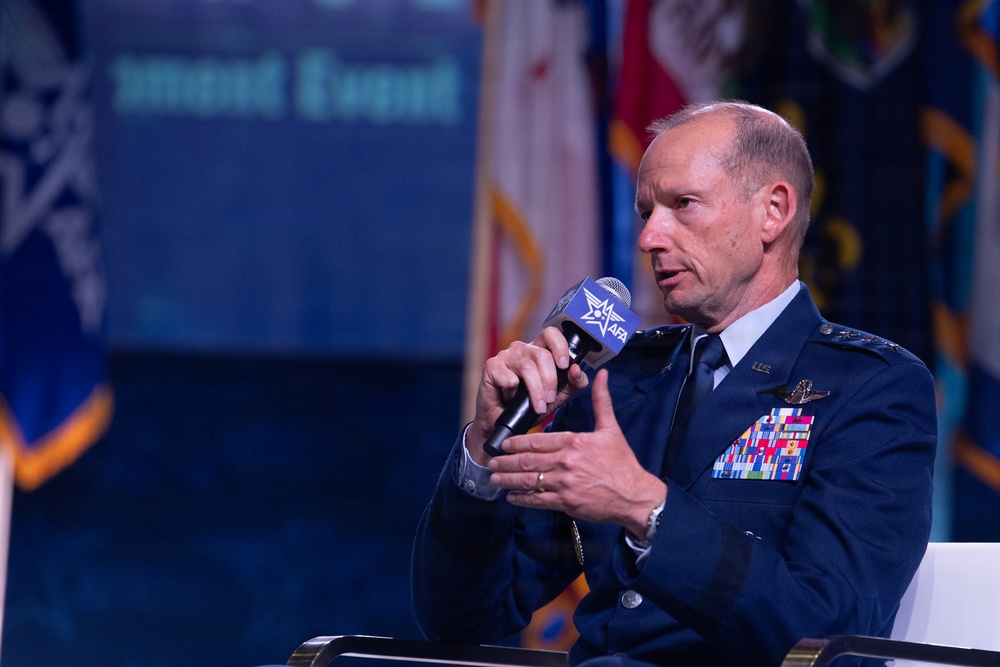 A senior U.S. Air Force officer in dress blue uniform is speaking into an AFA (Air Force Association) branded microphone at what appears to be an AFA conference or symposium. He is seated and gesturing with his free hand, suggesting an active panel discussion or interview. Multiple military flags and a blue branded backdrop are visible in the background.