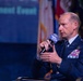 A senior U.S. Air Force officer in dress blue uniform is speaking into an AFA (Air Force Association) branded microphone at what appears to be an AFA conference or symposium. He is seated and gesturing with his free hand, suggesting an active panel discussion or interview. Multiple military flags and a blue branded backdrop are visible in the background.