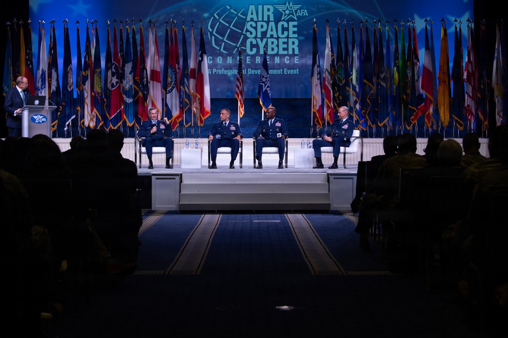 A panel discussion is taking place on stage at the AFA (Air Force Association) Air Space Cyber Conference, described as 'A Professional Development Event.' Four military officers in dress uniforms are seated on stage while a civilian moderator speaks at a podium on the left. The audience, visible in silhouette in the foreground, is seated in a darkened auditorium.