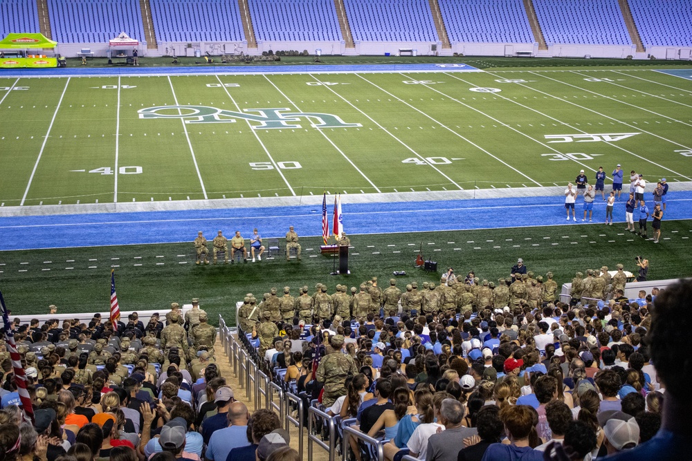 A military ceremony is taking place on the field of a college football stadium, with uniformed soldiers assembled in formation near a podium flanked by the American flag and what appears to be a state flag. A large crowd of civilians fills the lower seating sections, watching the proceedings. The stadium field markings and 'ACC' logo suggest this is an Atlantic Coast Conference college football venue.