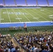 A military ceremony is taking place on the field of a college football stadium, with uniformed soldiers assembled in formation near a podium flanked by the American flag and what appears to be a state flag. A large crowd of civilians fills the lower seating sections, watching the proceedings. The stadium field markings and 'ACC' logo suggest this is an Atlantic Coast Conference college football venue.