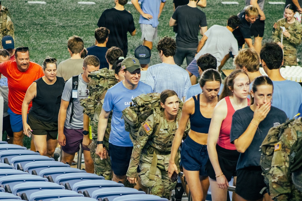 A large group of people are climbing stadium steps inside what appears to be an indoor or covered football stadium. The crowd includes both military personnel in camouflage uniforms carrying heavy rucksacks and civilians in athletic wear, suggesting a ruck march or fitness challenge event. The setting and mix of participants indicates this may be a military-civilian fitness or fundraising event.