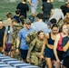 A large group of people are climbing stadium steps inside what appears to be an indoor or covered football stadium. The crowd includes both military personnel in camouflage uniforms carrying heavy rucksacks and civilians in athletic wear, suggesting a ruck march or fitness challenge event. The setting and mix of participants indicates this may be a military-civilian fitness or fundraising event.