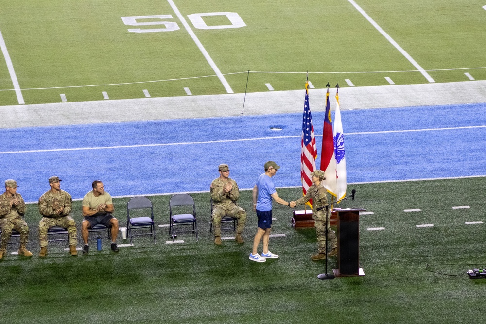 A military ceremony is taking place on the field of an indoor football stadium near the 50-yard line. A uniformed soldier is shaking hands with a civilian at a podium flanked by flags, while several other military personnel in camouflage uniforms are seated in folding chairs applauding. The setting suggests a formal recognition or award ceremony conducted during a sporting event.